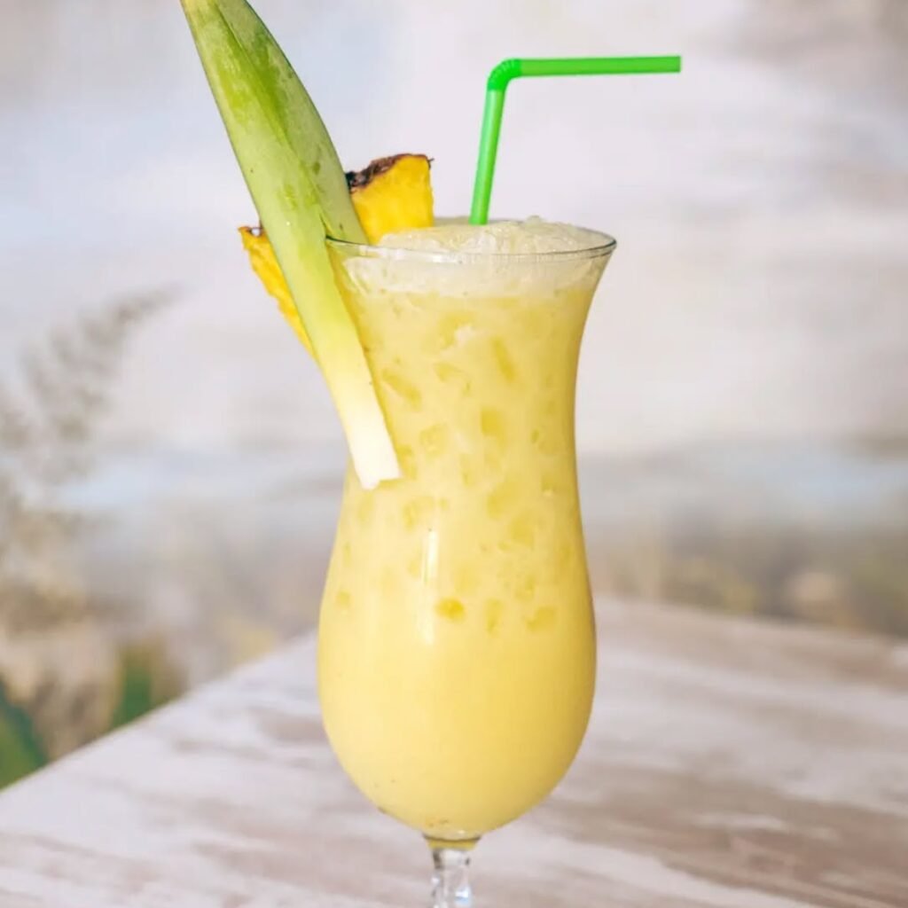 Cóctel Banana Colada de color amarillo claro, servido con pajita verde, decorado con una rodaja de piña y hoja tropical.