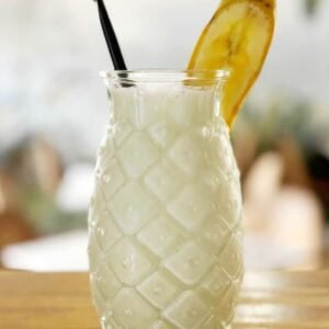 Batido de proteínas de color blanco servido en vaso de cristal tallado con pajita negra y rodaja de plátano deshidratado.