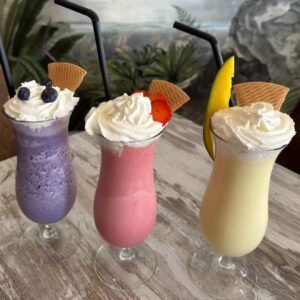 Tres batidos helados servidos en copas altas: uno morado con arándanos, otro rosa con fresa y el tercero amarillo con mango, todos decorados con nata, fruta y galleta.