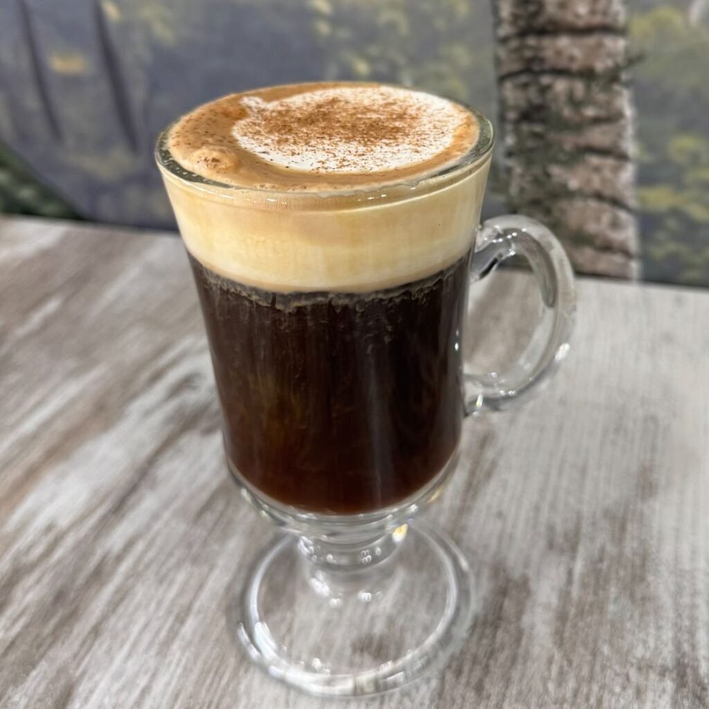 Taza de café Berni con capa de espuma de leche espolvoreada con canela, servida en vaso de cristal sobre mesa de madera