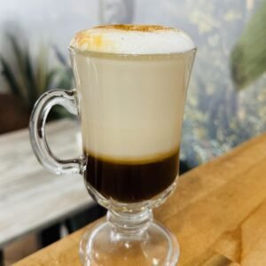 Cappuccino con abundante espuma servido en taza de cristal, decorado con un toque de canela