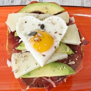 Tostada gourmet con aguacate, jamón, queso y huevo en forma de corazón