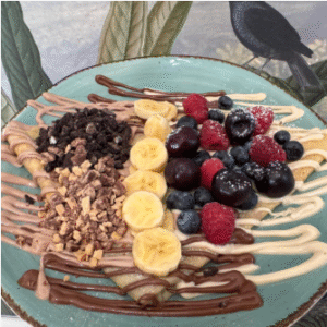 Crêpe decorado con sirope de chocolate y vainilla, frutas del bosque, plátano, virutas de chocolate y frutos secos sobre plato azul
