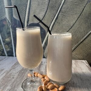 Batido natural de avena, leche de coco, plátano y almendras servido en vaso y copa sobre mesa de madera