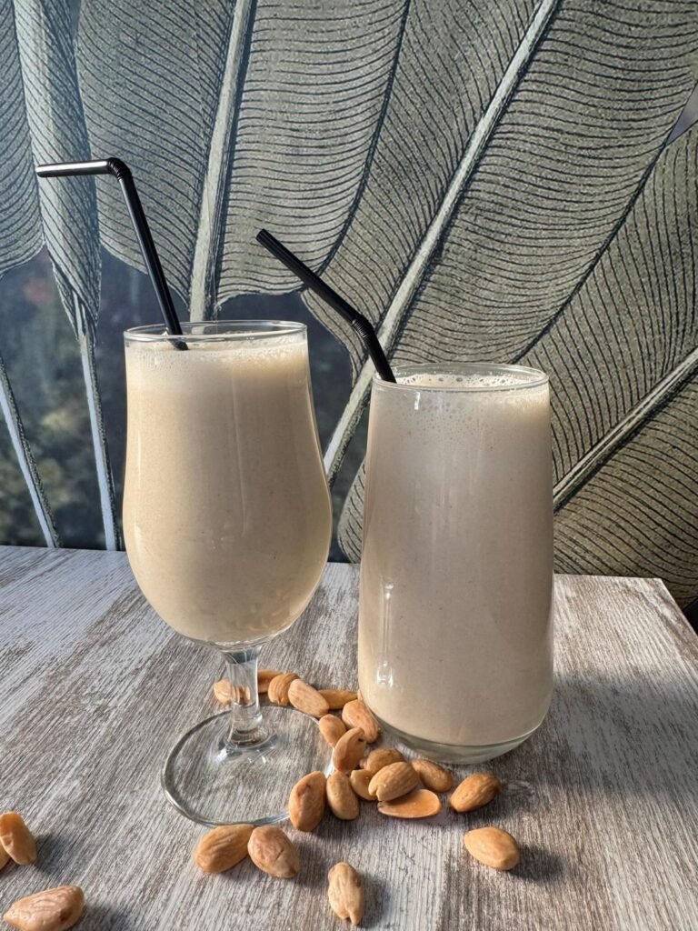 Batido natural de avena, leche de coco, plátano y almendras servido en vaso y copa sobre mesa de madera